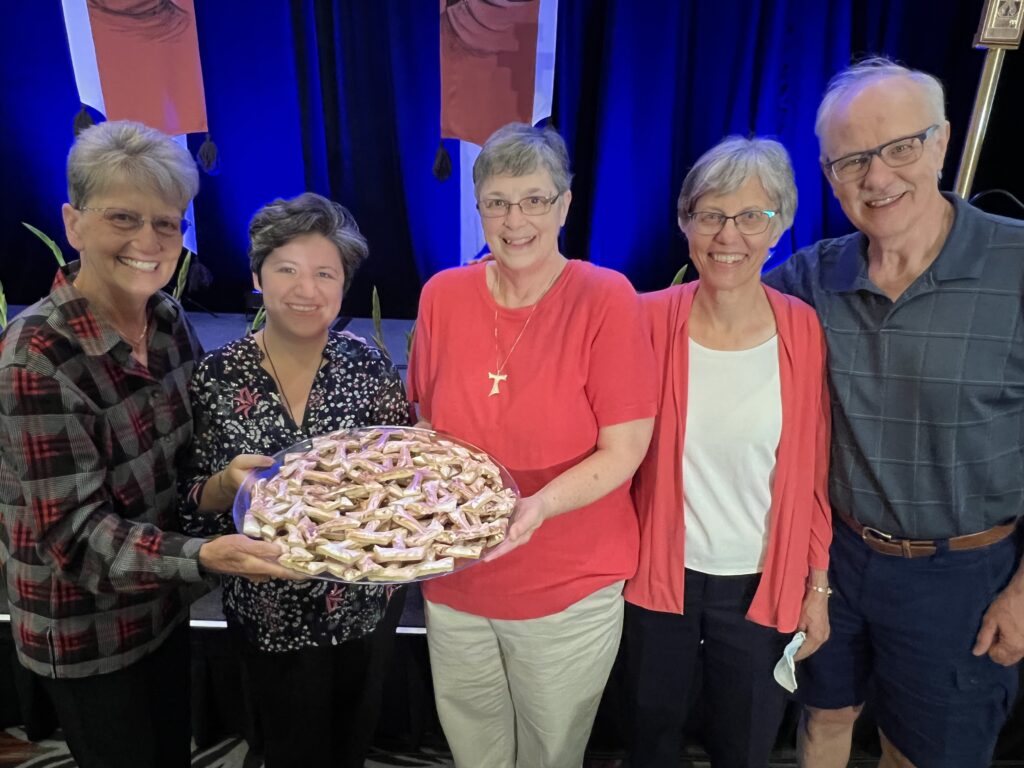 Five Federation members smile as together they hold a large plate of cookies in the shape of the Tau cross.